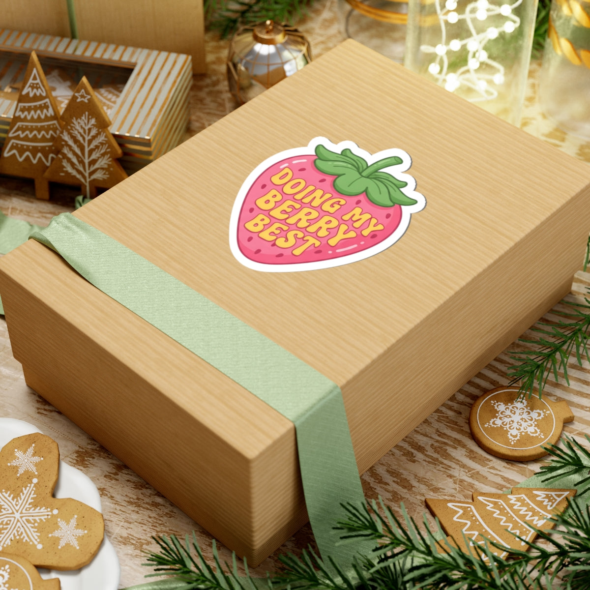 A square kraft paper gift box wrapped with a pale green ribbon and topped with a strawberry-shaped sticker that reads 'DOING MY BERRY BEST,' styled on a tabletop with holiday cookies, greenery, and gift boxes in the background.