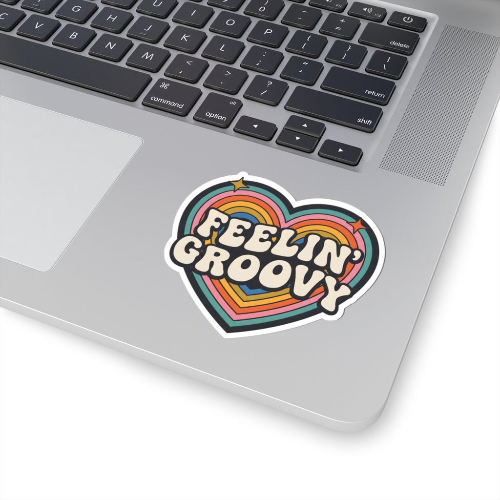 A heart-shaped retro sticker with the words 'FEELIN' GROOVY' in white bubble letters over rainbow stripes, shown applied to the silver palm rest of a laptop keyboard.
