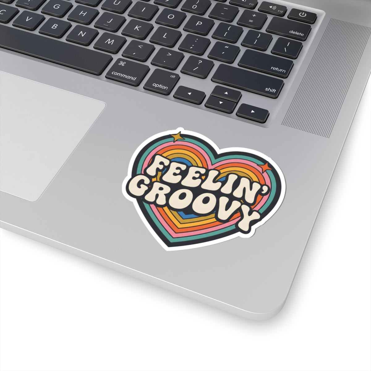 A heart-shaped retro sticker with the words 'FEELIN' GROOVY' in white bubble letters over rainbow stripes, shown applied to the silver palm rest of a laptop keyboard.