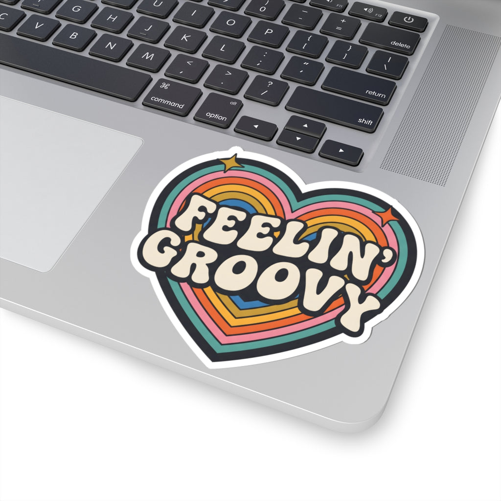 A heart-shaped vinyl sticker with the words 'FEELIN' GROOVY' in retro cream lettering over rainbow stripes, shown applied to the palm rest of a silver laptop.