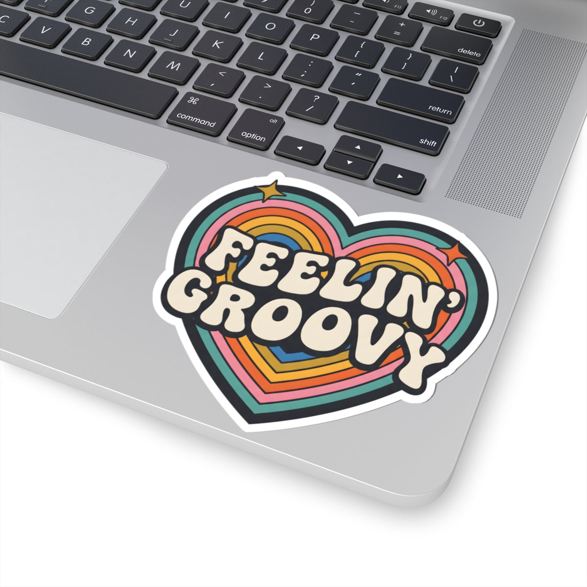 A heart-shaped vinyl sticker with the words 'FEELIN' GROOVY' in retro cream lettering over rainbow stripes, shown applied to the palm rest of a silver laptop.
