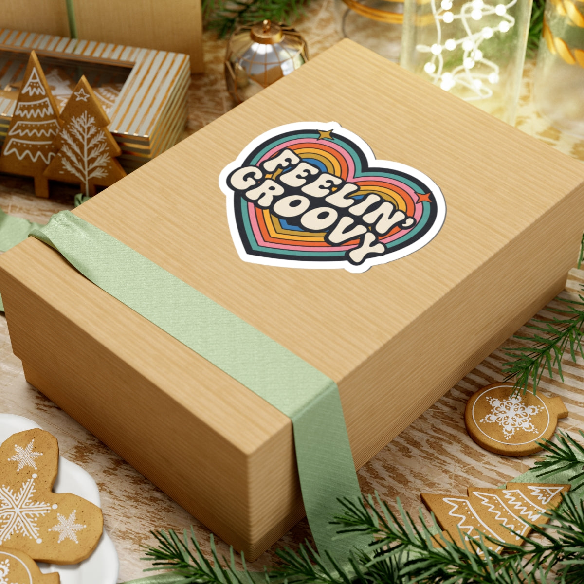 A kraft paper gift box tied with a pale green ribbon, featuring a heart-shaped retro rainbow sticker that reads 'FEELIN' GROOVY.' The box is styled on a tabletop with holiday decorations, including gingerbread cookies, evergreen branches, and glassware.
