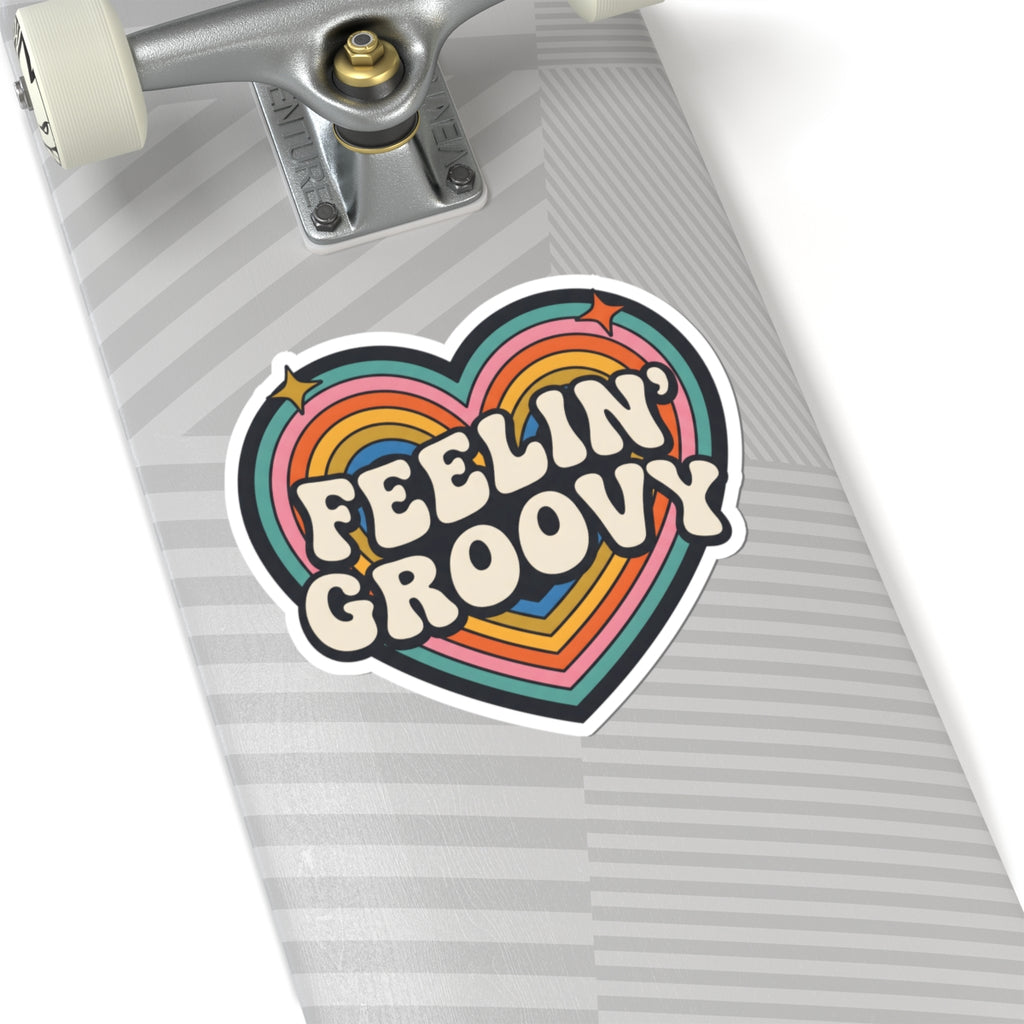 A heart-shaped sticker with the words 'FEELIN' GROOVY' in retro cream lettering, layered over rainbow stripes in pink, orange, yellow, green, blue, and purple, shown applied to the deck of a skateboard.