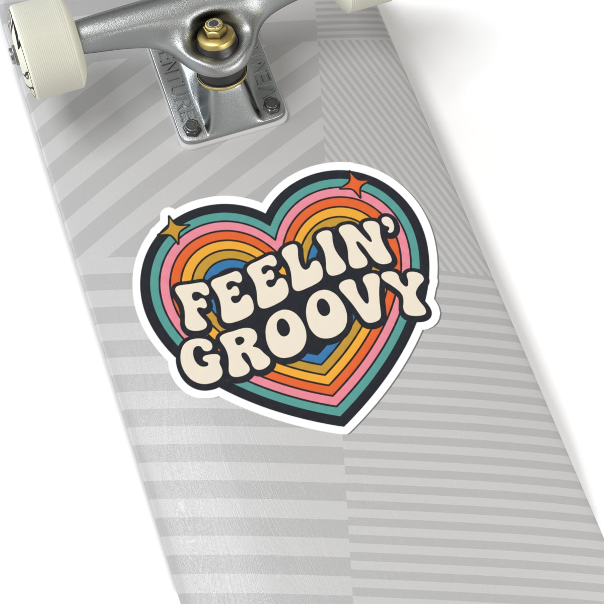 A heart-shaped sticker with the words 'FEELIN' GROOVY' in retro cream lettering, layered over rainbow stripes in pink, orange, yellow, green, blue, and purple, shown applied to the deck of a skateboard.