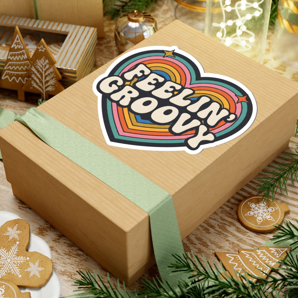 A kraft paper gift box tied with a pale green ribbon, featuring a colorful heart-shaped sticker that reads “FEELIN’ GROOVY,” styled on a rustic tabletop with holiday cookies, pine branches, and warm ambient decor.