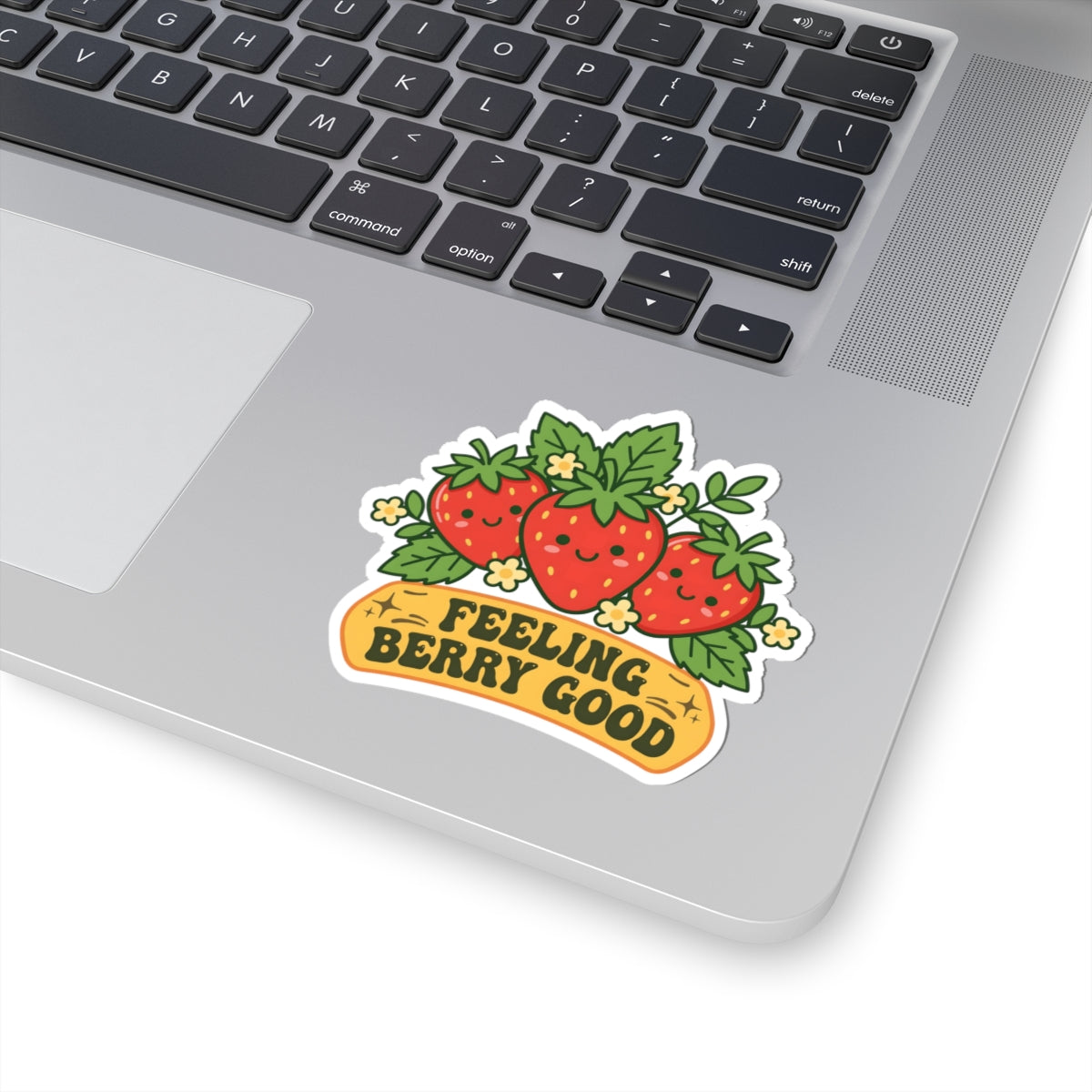 A kiss-cut vinyl sticker featuring three smiling red strawberries with green leaves and the text 'FEELING BERRY GOOD' on a yellow banner, shown applied to the silver palm rest of a laptop keyboard.