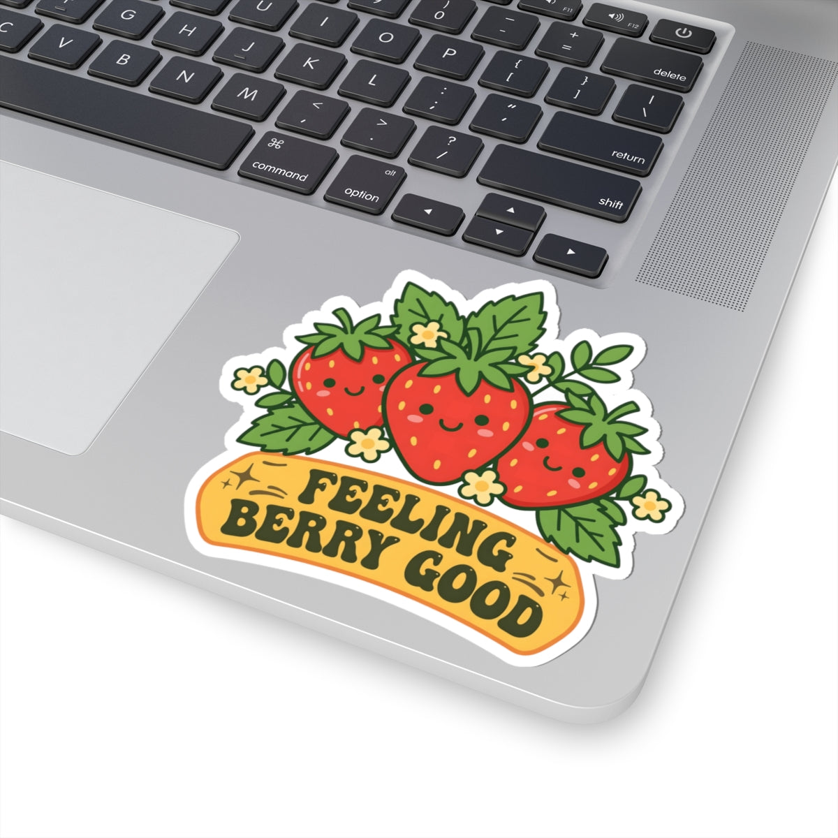 A kiss-cut vinyl sticker featuring three smiling cartoon strawberries with green leaves and small yellow flowers, above a yellow banner that reads “FEELING BERRY GOOD.” The sticker is shown applied to the corner of a silver laptop keyboard.