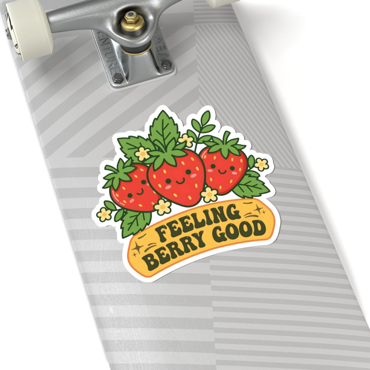 A close-up of a skateboard deck with a colorful sticker featuring three smiling strawberries and the text 'FEELING BERRY GOOD'. The skateboard has a gray and white striped pattern, and the truck is visible at the top.