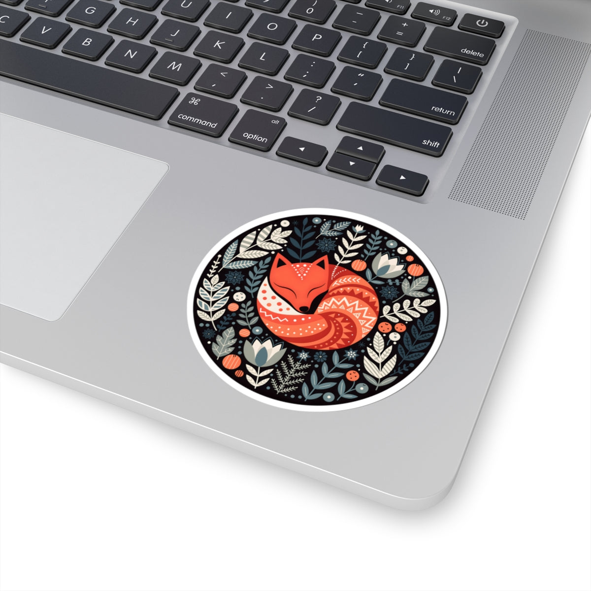 A round sticker with a whimsical fox and floral design, shown applied to the palm rest of a silver laptop.