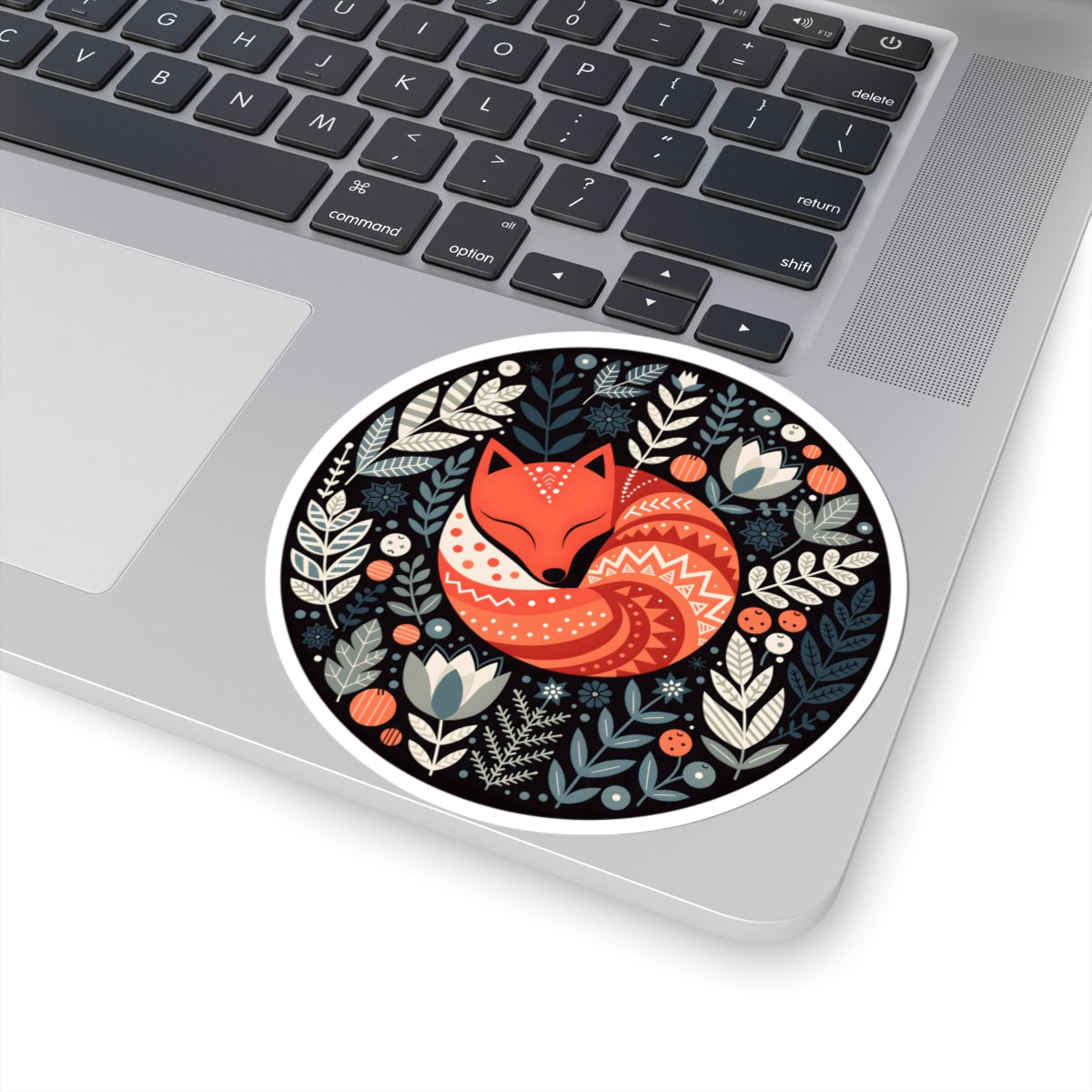A round vinyl sticker featuring a stylized orange fox with white and red patterns, surrounded by a dark blue and beige botanical design, shown applied to the corner of a silver laptop.
