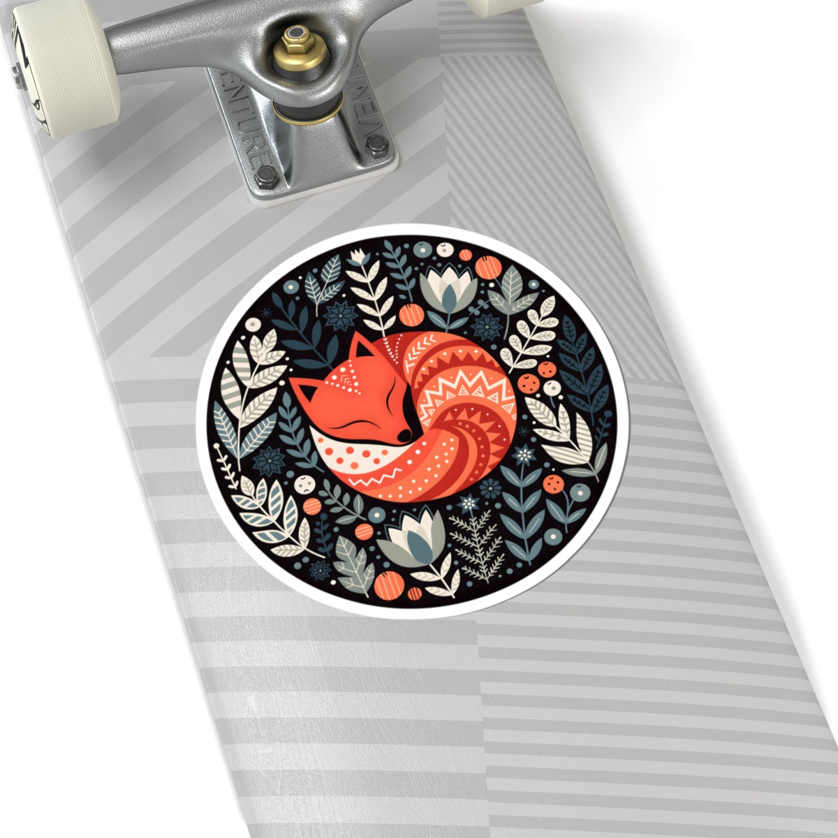 A circular sticker featuring a stylized orange fox surrounded by botanical illustrations, shown applied to the deck of a skateboard with a gray striped pattern.