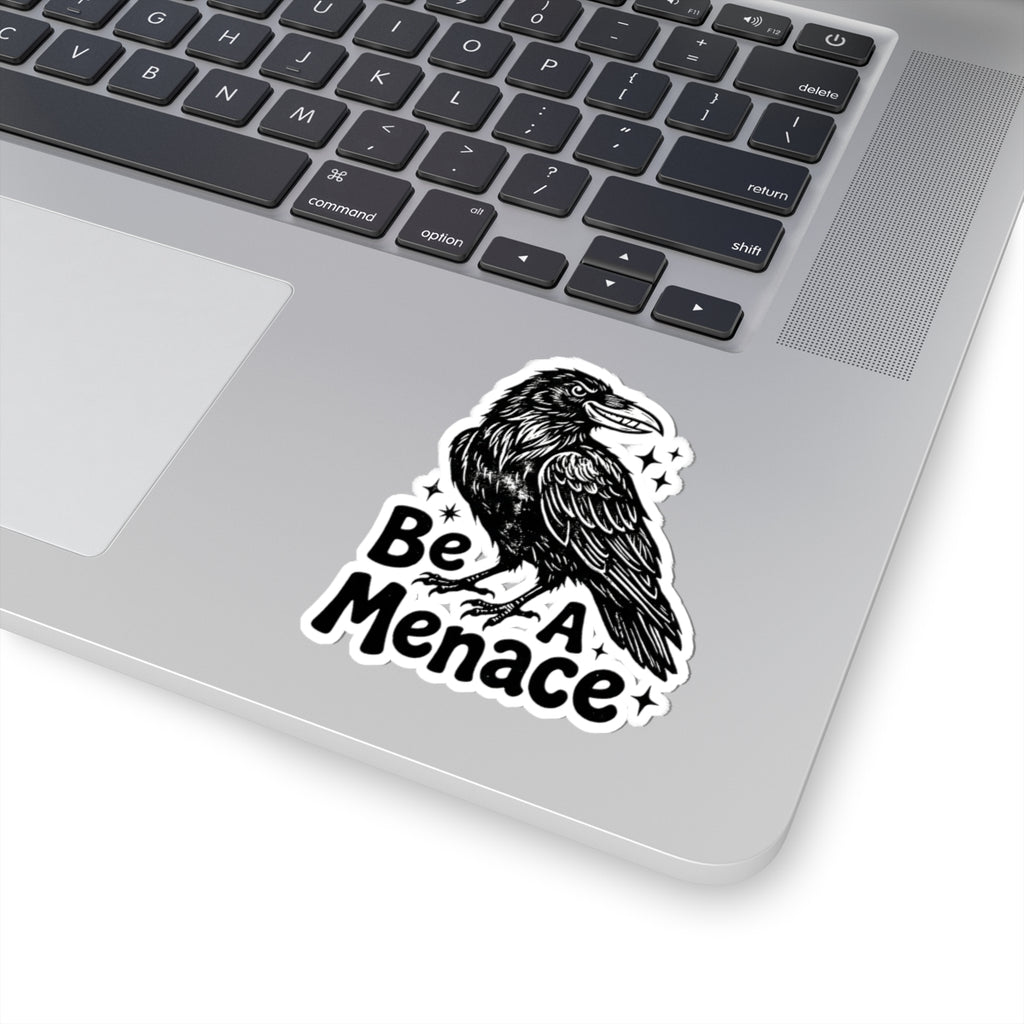 A kiss-cut vinyl sticker featuring a black raven illustration and the text 'Be A Menace' with small star accents, shown applied to the lower corner of a silver laptop keyboard.