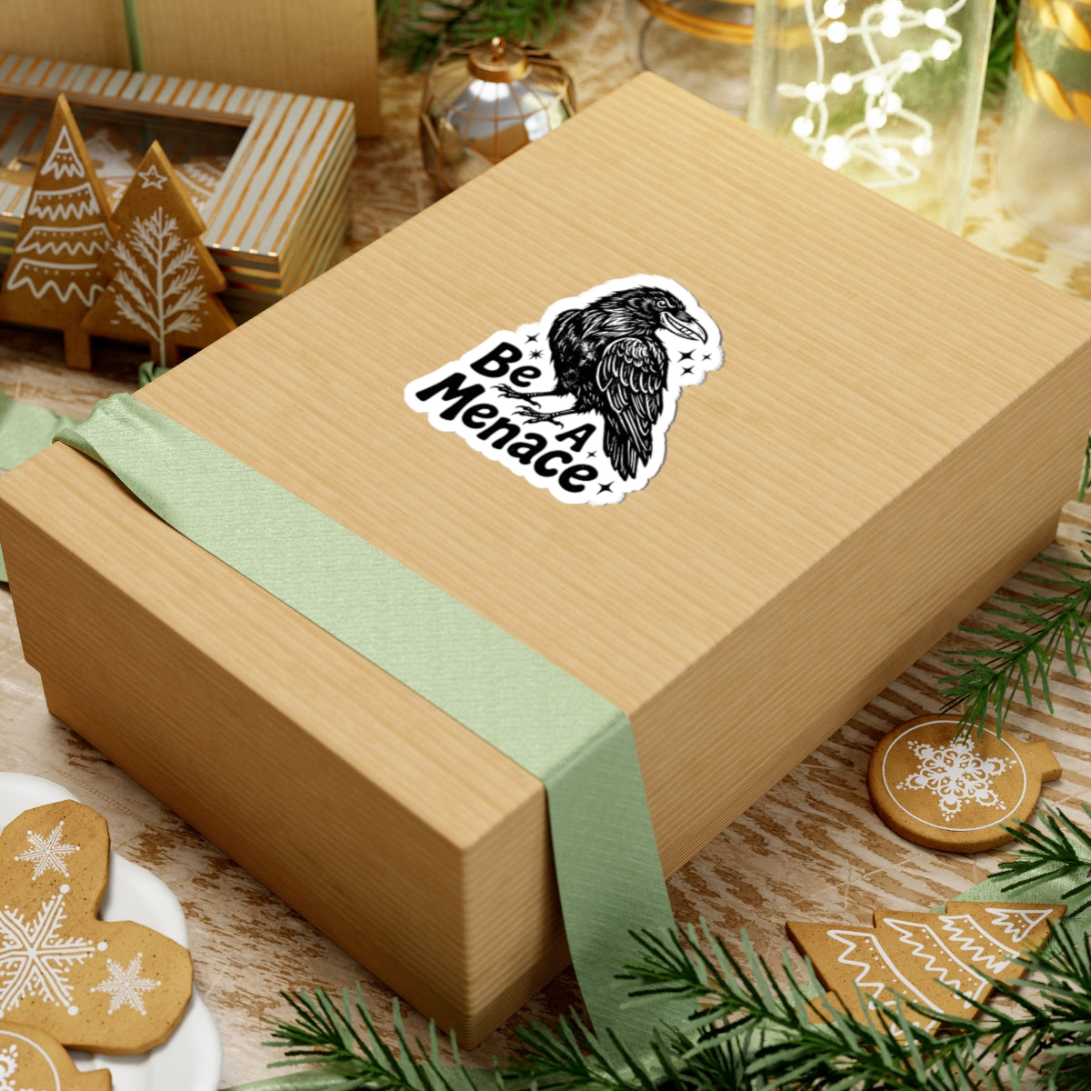 A kraft paper gift box tied with a pale green ribbon, featuring a kiss-cut sticker of a black raven and the words 'Be A Menace' on the top surface. The box is styled on a rustic wooden surface with Christmas cookies, evergreen branches, and holiday decorations.