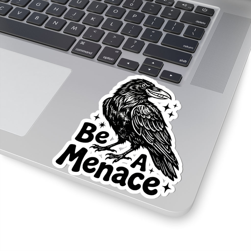 A kiss-cut vinyl sticker featuring a black and white illustration of a raven with the text 'Be A Menace' and small star accents, shown applied to the corner of a silver laptop.