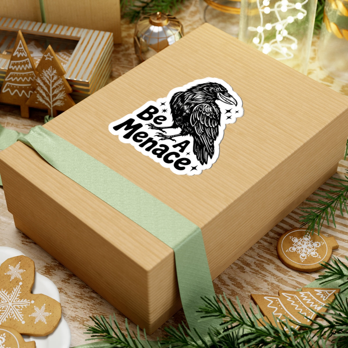 A kraft paper gift box tied with a pale green ribbon, featuring a kiss-cut sticker of a black raven and the words 'Be A Menace' on top, styled on a wooden surface with holiday decorations.