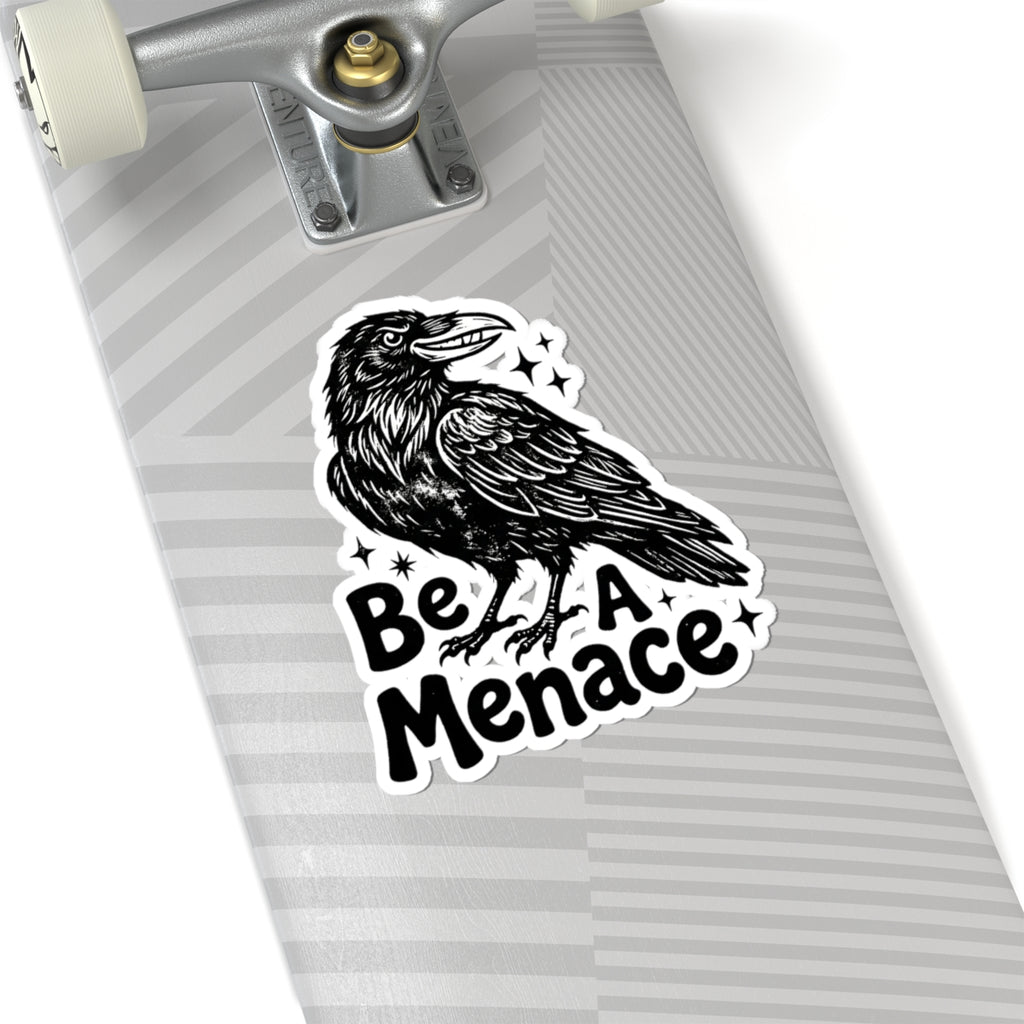 A kiss-cut sticker featuring a black raven illustration and the text 'Be A Menace' with small star accents, shown applied to the side of a gray skateboard deck.