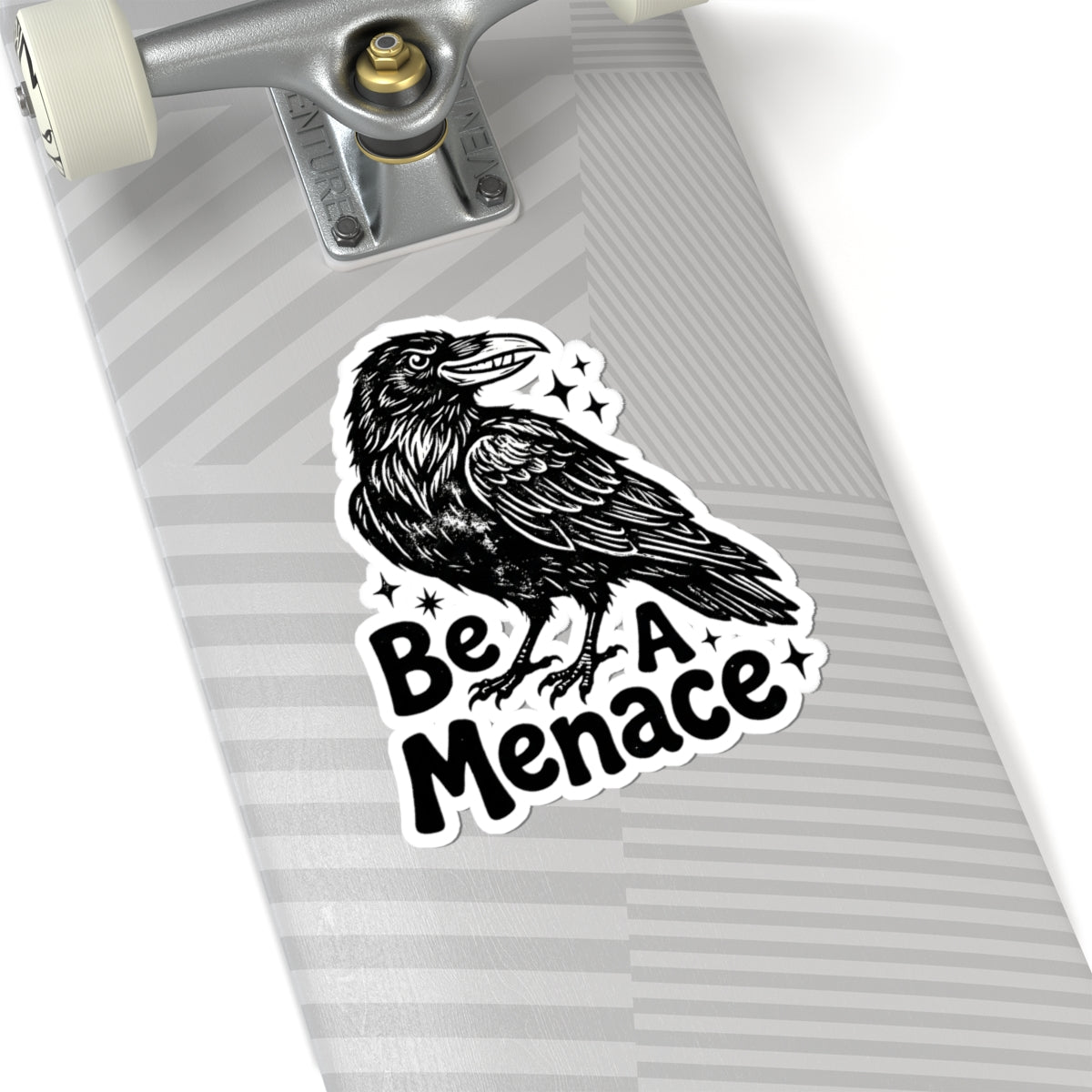 A kiss-cut sticker featuring a black raven illustration and the text 'Be A Menace' with small star accents, shown applied to the side of a gray skateboard deck.