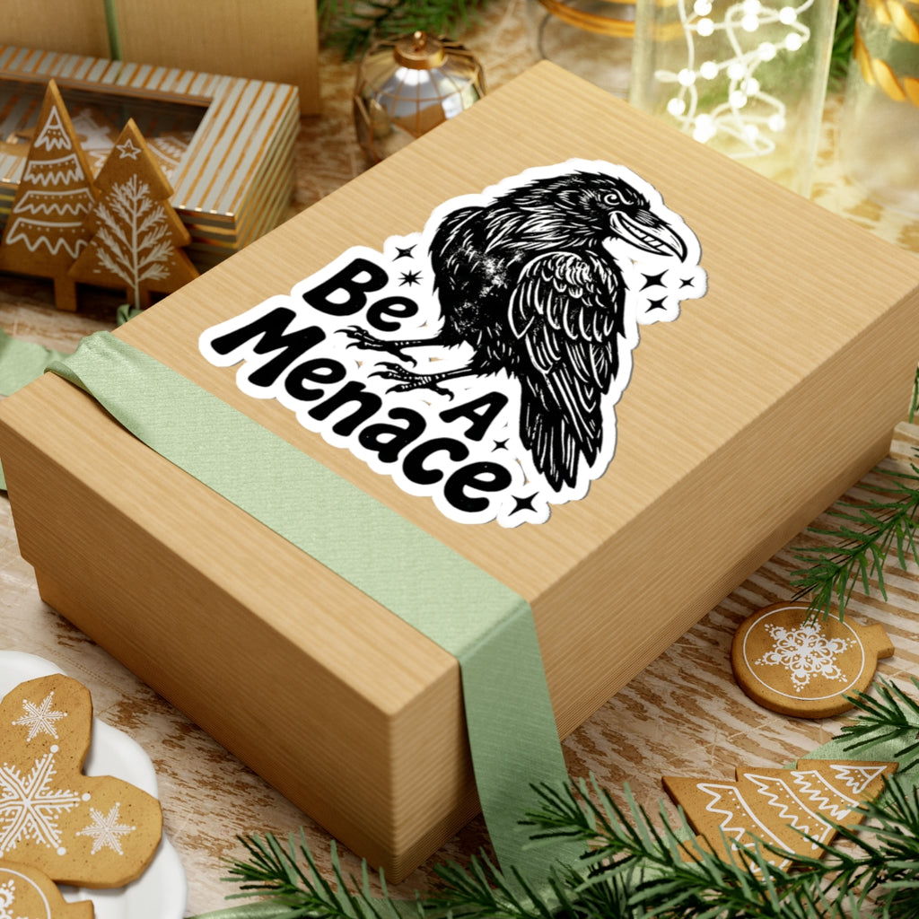 A kraft paper gift box wrapped with a pale green ribbon, featuring a kiss-cut sticker of a black raven and the words “Be A Menace.” The box is styled on a rustic wooden surface with holiday decorations including gingerbread cookies, evergreen branches, and a small decorative box in the background.
