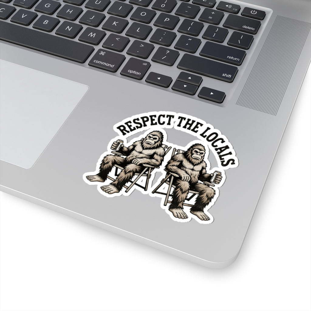 A kiss-cut vinyl sticker applied to the silver palm rest of a laptop keyboard. The sticker features a black and white graphic of two Bigfoot figures sitting in chairs, with the text 'RESPECT THE LOCALS' arched above them.