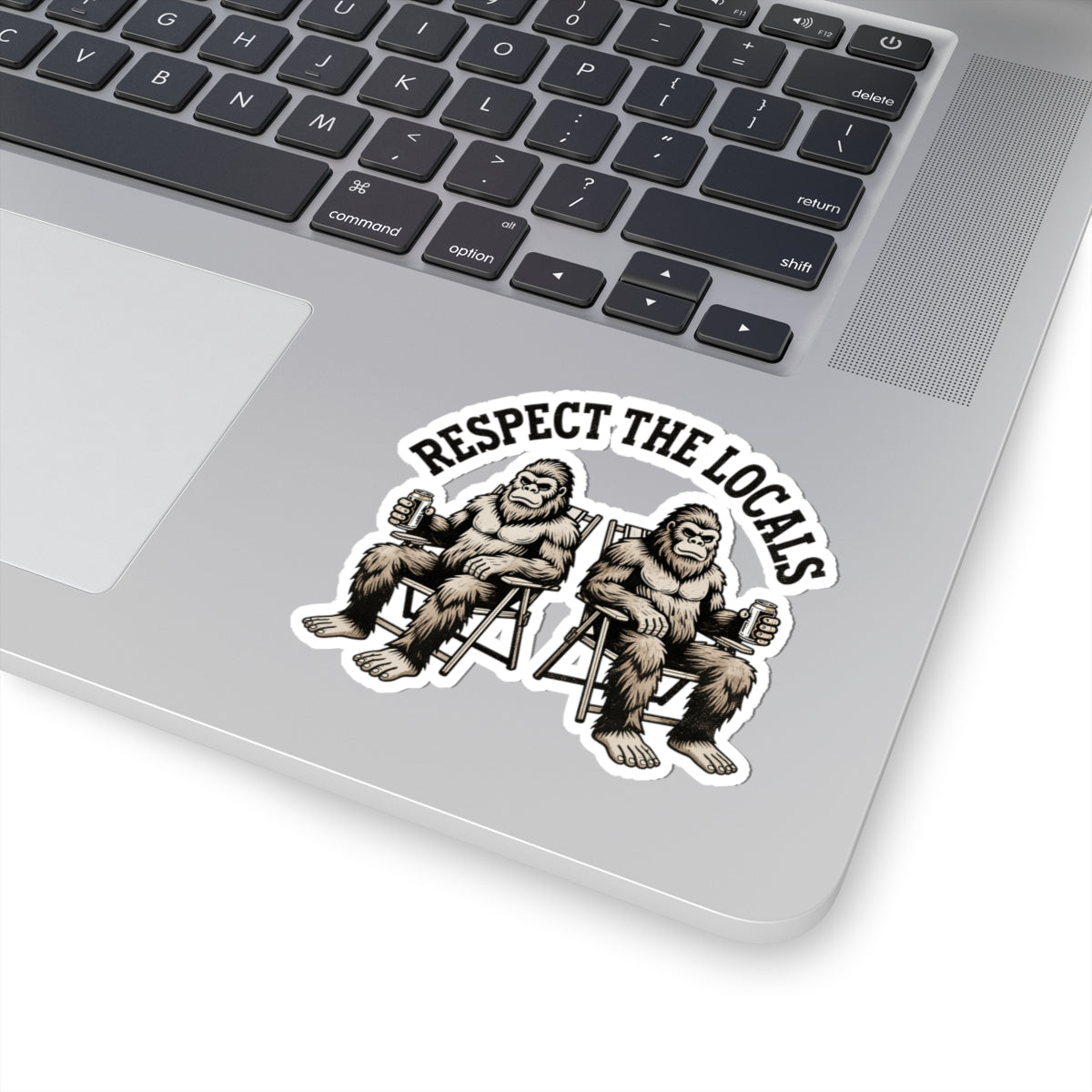 A kiss-cut vinyl sticker applied to the silver palm rest of a laptop keyboard. The sticker features a black and white graphic of two Bigfoot figures sitting in chairs, with the text 'RESPECT THE LOCALS' arched above them.