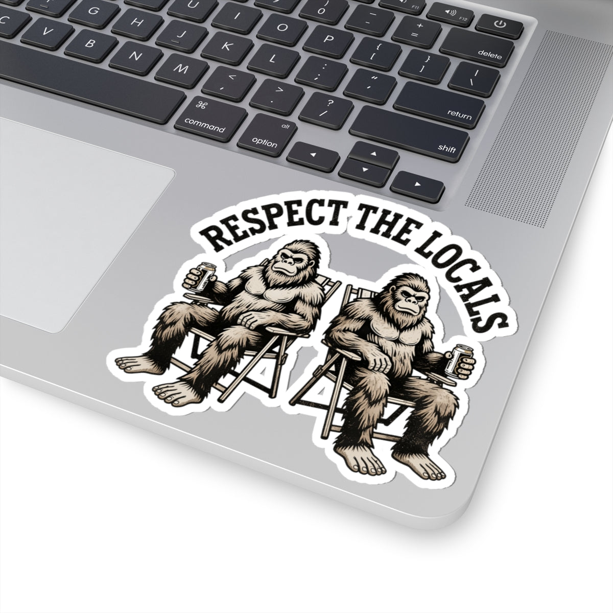 A kiss-cut vinyl sticker applied to the corner of a silver laptop. The sticker features an illustration of two Bigfoot figures sitting in chairs holding beer mugs under the text 'RESPECT THE LOCALES'.

