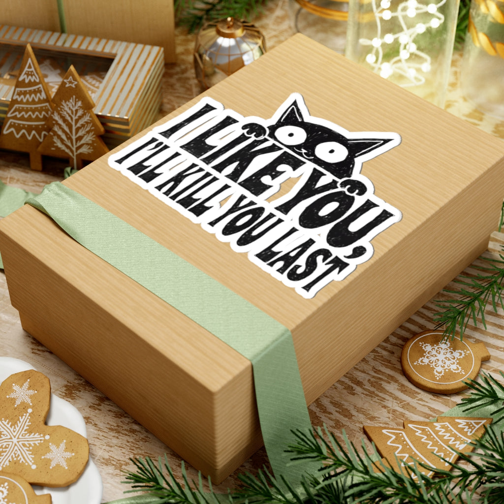 A square kraft paper gift box wrapped with a pale green ribbon, featuring a kiss-cut sticker on top with a black cat face and the text “I LIKE YOU, I'LL KILL YOU LAST.” The box is styled on a rustic wooden surface with holiday decorations including gingerbread cookies, evergreen branches, and another gift box in the background.