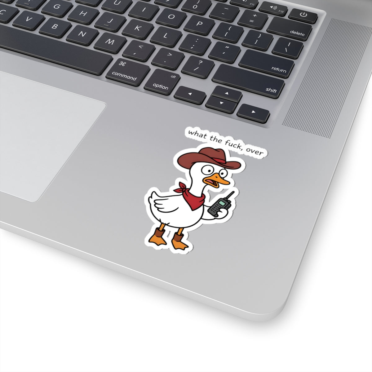 A cartoon duck sticker with the text 'what the fuck, over' is shown on the palm rest of a silver laptop. The sticker features a white duck wearing a brown cowboy hat and red bandana, holding a walkie-talkie.