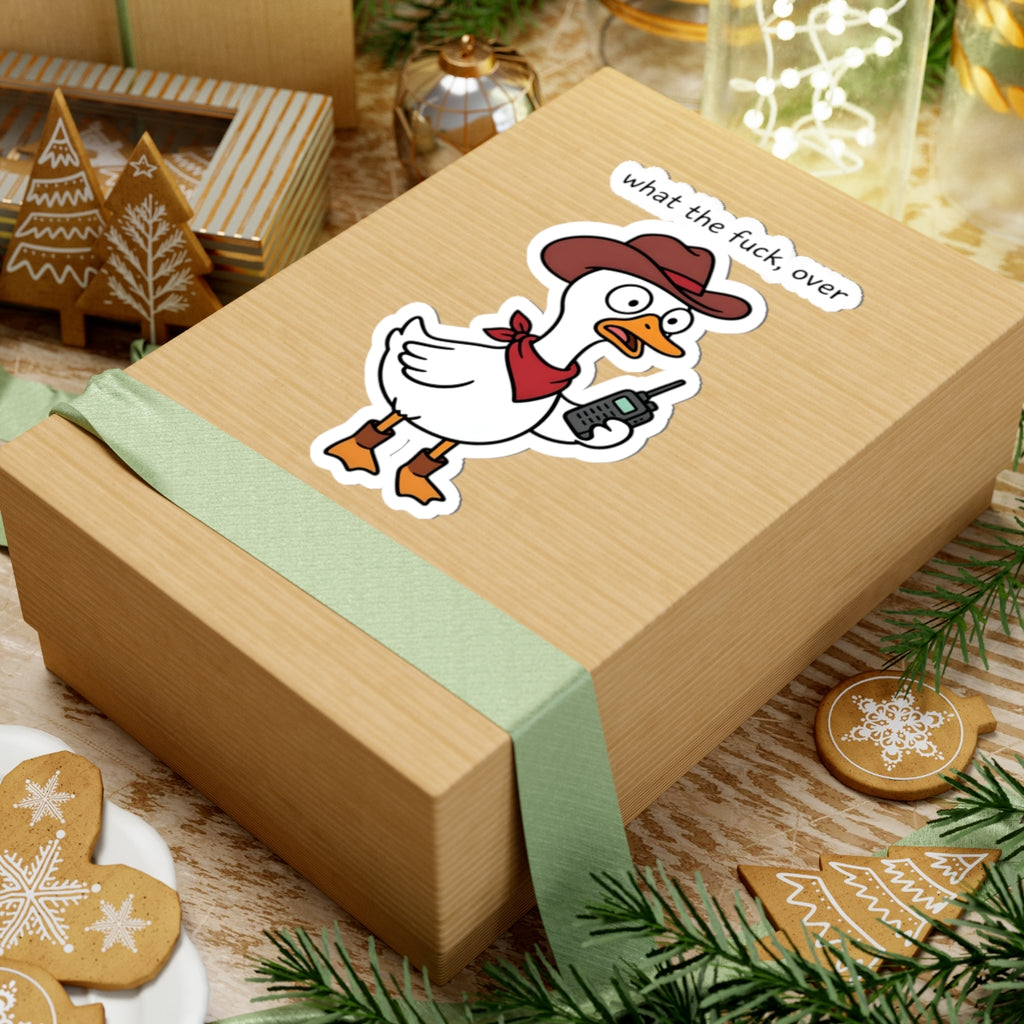 A light brown kraft paper gift box tied with a pale green ribbon, featuring a cartoon duck sticker wearing a cowboy hat and the text 'what the fuck over'. The box is styled on a wooden surface with holiday cookies, greenery, and warm background lights.
