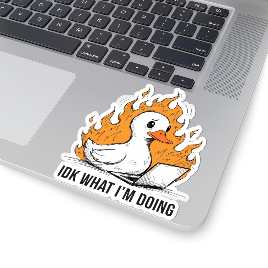 A kiss-cut vinyl sticker featuring a white duck surrounded by orange flames, with the text 'IDK WHAT I'M DOING' below, shown applied to the corner of a silver laptop.