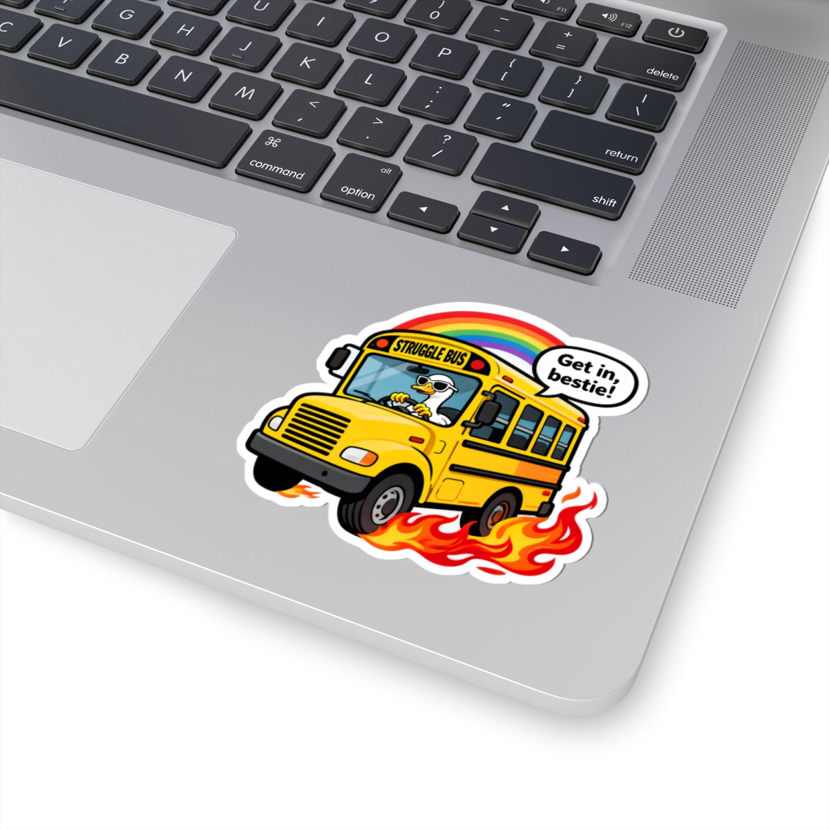 A kiss-cut vinyl sticker of a cartoon yellow school bus with flames and a rainbow, applied to the palm rest of a silver laptop. The sticker features a speech bubble that reads 'Get in, bestie!'.