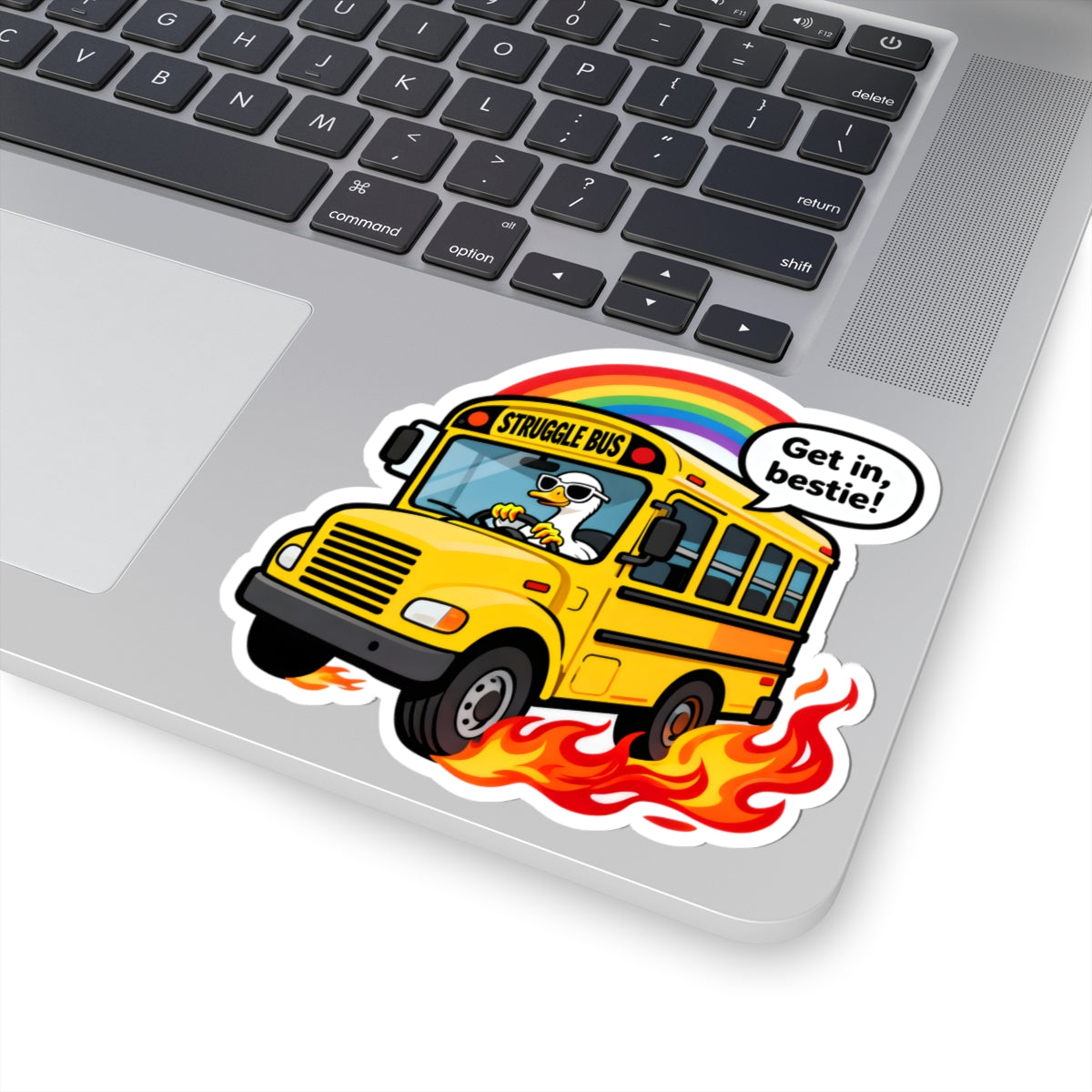 A kiss-cut vinyl sticker applied to the corner of a silver laptop. The sticker features a cartoon yellow school bus with flames, a rainbow, a dog driver, and a speech bubble that reads 'Get in, bestie!'.