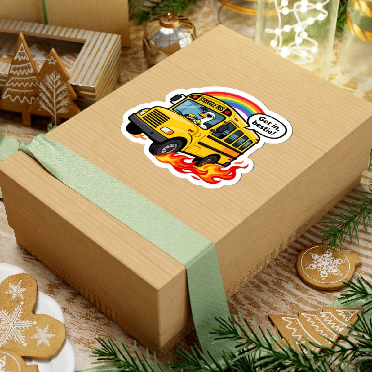 A kraft paper gift box tied with a pale green ribbon, decorated with a kiss-cut sticker of a yellow school bus with flames and a rainbow. The box is styled on a rustic wooden surface with Christmas cookies, evergreen branches, and holiday decorations.