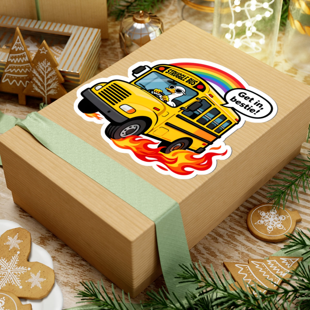 A square kraft gift box wrapped with a pale green ribbon, featuring a colorful kiss-cut sticker of a yellow school bus with flames and a rainbow. The box is styled on a wooden surface with holiday cookies, evergreen branches, and other gift items in the background.