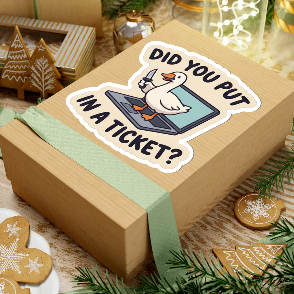 A light brown kraft gift box wrapped with a pale green ribbon, featuring a kiss-cut sticker that reads 'DID YOU PUT IN A TICKET?' with a cartoon goose on a laptop. The box is styled on a rustic wooden surface with Christmas cookies, evergreen branches, and warm holiday decor in the background.