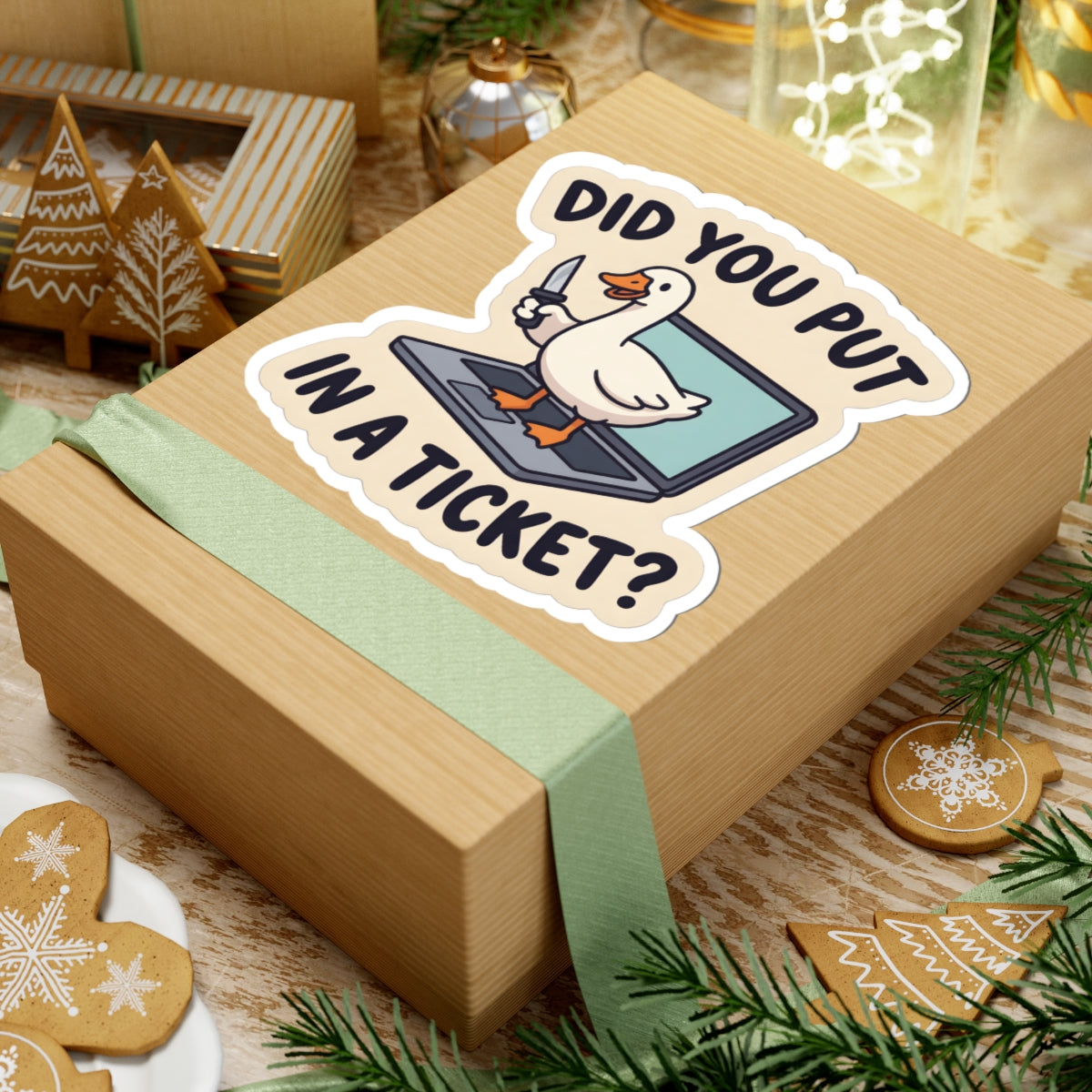 A light brown kraft gift box wrapped with a pale green ribbon, featuring a kiss-cut sticker that reads 'DID YOU PUT IN A TICKET?' with a cartoon goose on a laptop. The box is styled on a rustic wooden surface with Christmas cookies, evergreen branches, and warm holiday decor in the background.
