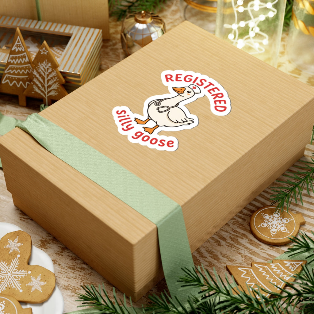 A light brown kraft gift box tied with a pale green ribbon, featuring a kiss-cut sticker of a white goose with the words 'REGISTERED silly goose' on top. The box is styled on a rustic wooden surface with holiday decorations including gingerbread cookies, pine branches, and a small gift box in the background.