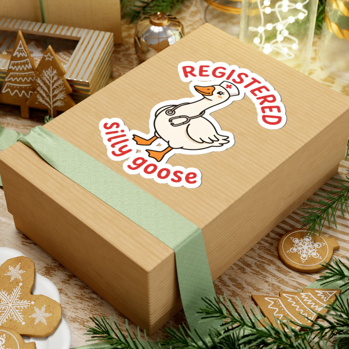 A square kraft gift box wrapped with a pale green ribbon, featuring a kiss-cut sticker of a white goose with orange feet and the words “REGISTERED silly goose” in red and white. The box is styled on a rustic wooden surface with gingerbread cookies, evergreen branches, and holiday decor.