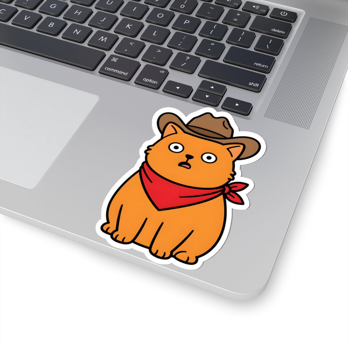 A kiss-cut vinyl sticker of an orange cartoon cat wearing a brown cowboy hat and red bandana, shown applied to the silver surface of a laptop next to the keyboard.