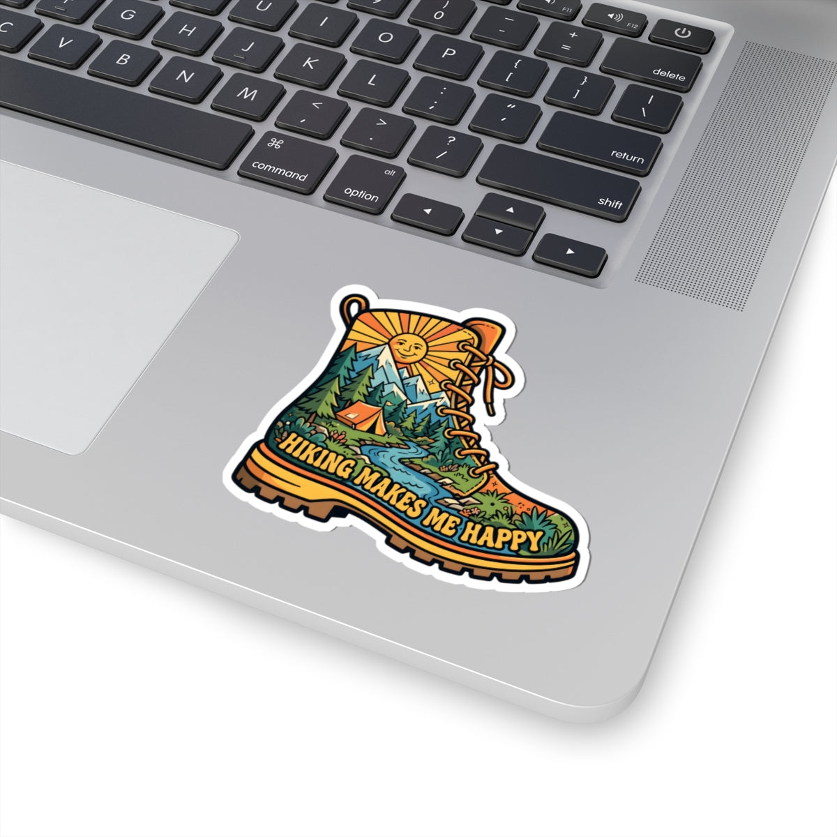 A kiss-cut vinyl sticker of a hiking boot with a scenic landscape and the text 'HIKING MAKES ME HAPPY' applied to the palm rest of a silver laptop.