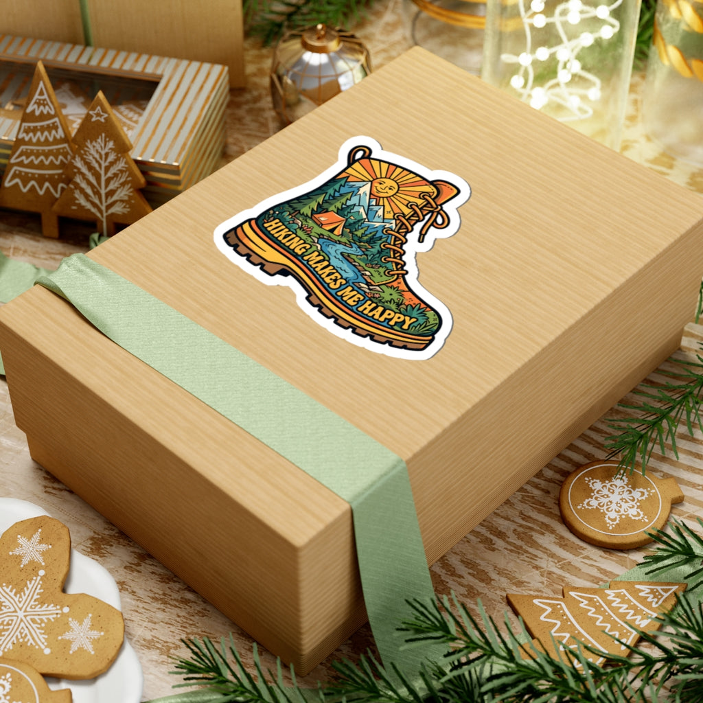 A light brown kraft gift box tied with a pale green ribbon, featuring a kiss-cut sticker of a hiking boot with a mountain landscape and the words 'HIKING MAKES ME HAPPY'. The box is styled on a rustic wooden surface with Christmas cookies, pine branches, and holiday decorations.
