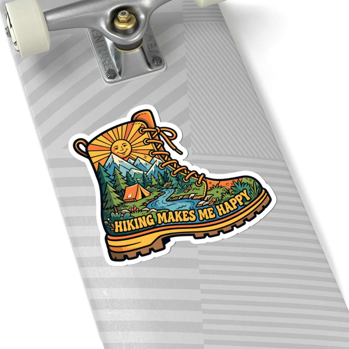A kiss-cut sticker shaped like a hiking boot, shown on the deck of a skateboard. The sticker features a colorful illustration of a mountain landscape with a tent, trees, and a river, with the text 'HIKING MAKES ME HAPPY' in yellow.
