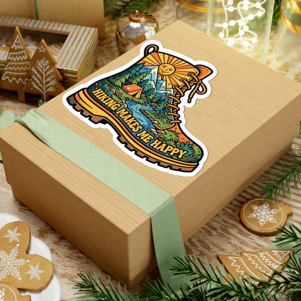 A square kraft gift box wrapped with a pale green ribbon, topped with a kiss-cut sticker shaped like a hiking boot. The sticker features a colorful scenic landscape and the phrase 'HIKING MAKES ME HAPPY.