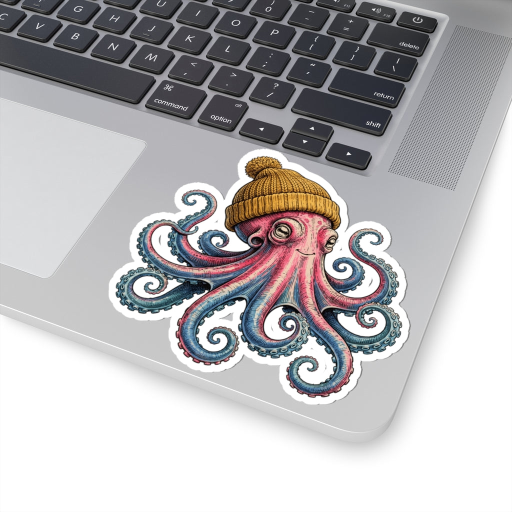 A kiss-cut vinyl sticker of a pink and blue octopus wearing a yellow beanie, shown applied to the silver surface of a laptop near the keyboard.