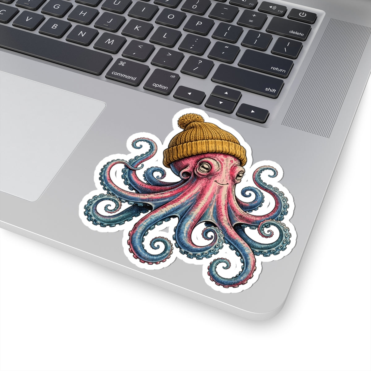 A kiss-cut vinyl sticker of a pink and blue octopus wearing a yellow beanie, shown applied to the silver surface of a laptop near the keyboard.