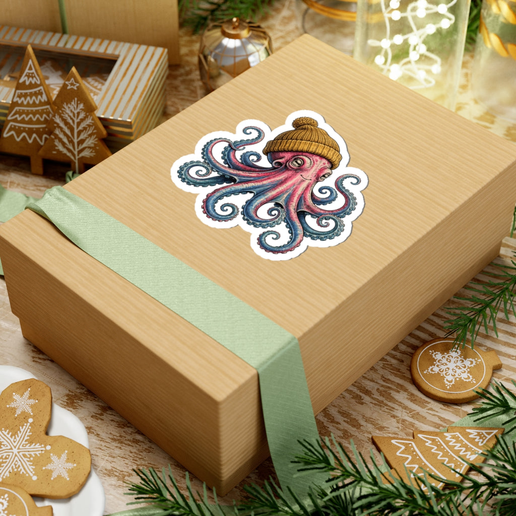 A kraft paper gift box wrapped with a pale green ribbon, decorated with a colorful octopus sticker wearing a knit beanie. The box is styled on a rustic wooden surface with Christmas cookies, pine branches, and holiday decorations.