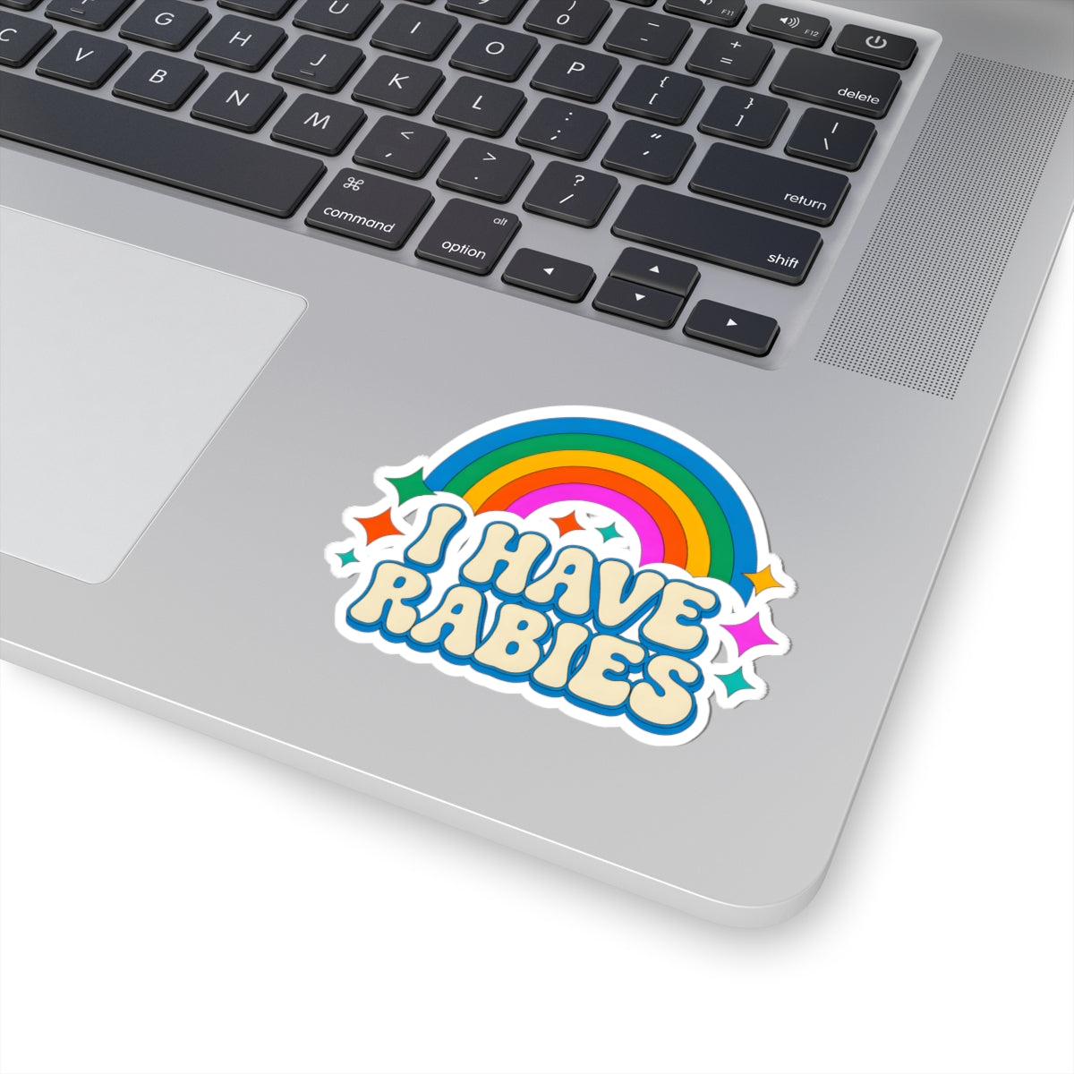 A colorful vinyl sticker reading 'I HAVE RABIES' in bubbly blue letters with a rainbow arch and stars, shown applied to the silver palm rest of a laptop keyboard.