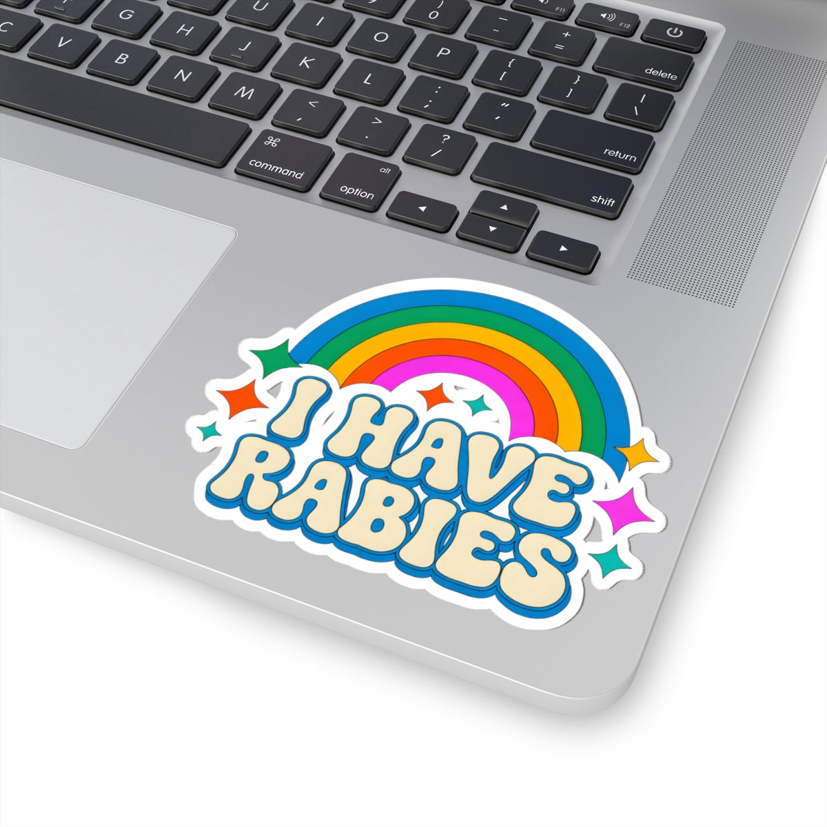 A kiss-cut vinyl sticker with the text 'I HAVE RABIES' in bubbly cream lettering outlined in blue, placed on the corner of a silver laptop next to the keyboard. The sticker features a vibrant rainbow arch and small colorful stars.