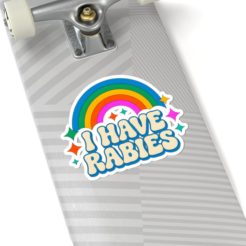 A kiss-cut vinyl sticker reading 'I HAVE RABIES' in a playful retro font, featuring a rainbow and stars, shown applied to the deck of a skateboard.
