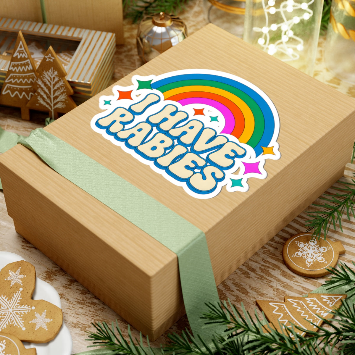 A square kraft gift box wrapped with a pale green ribbon, featuring a colorful die-cut sticker that reads “I HAVE RABIES” beneath a rainbow and stars. The box is styled on a rustic wooden surface with holiday cookies, evergreen branches, and other gift items in the background.