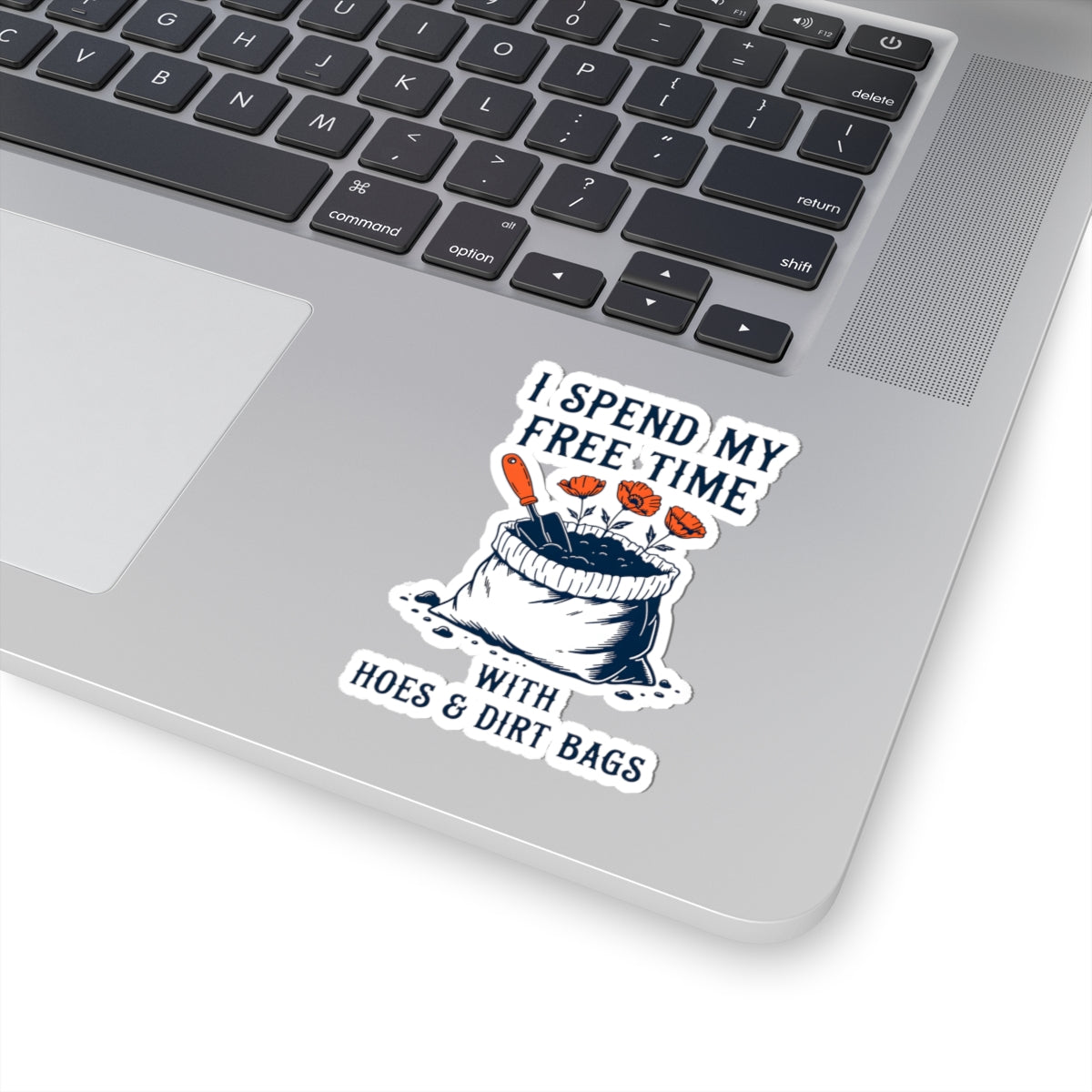 A kiss-cut vinyl sticker applied to the lower right corner of a silver laptop. The sticker features a cartoon illustration of garden supplies, with the text 'I SPEND MY FREE TIME WITH HOES & DIRT BAGS'.